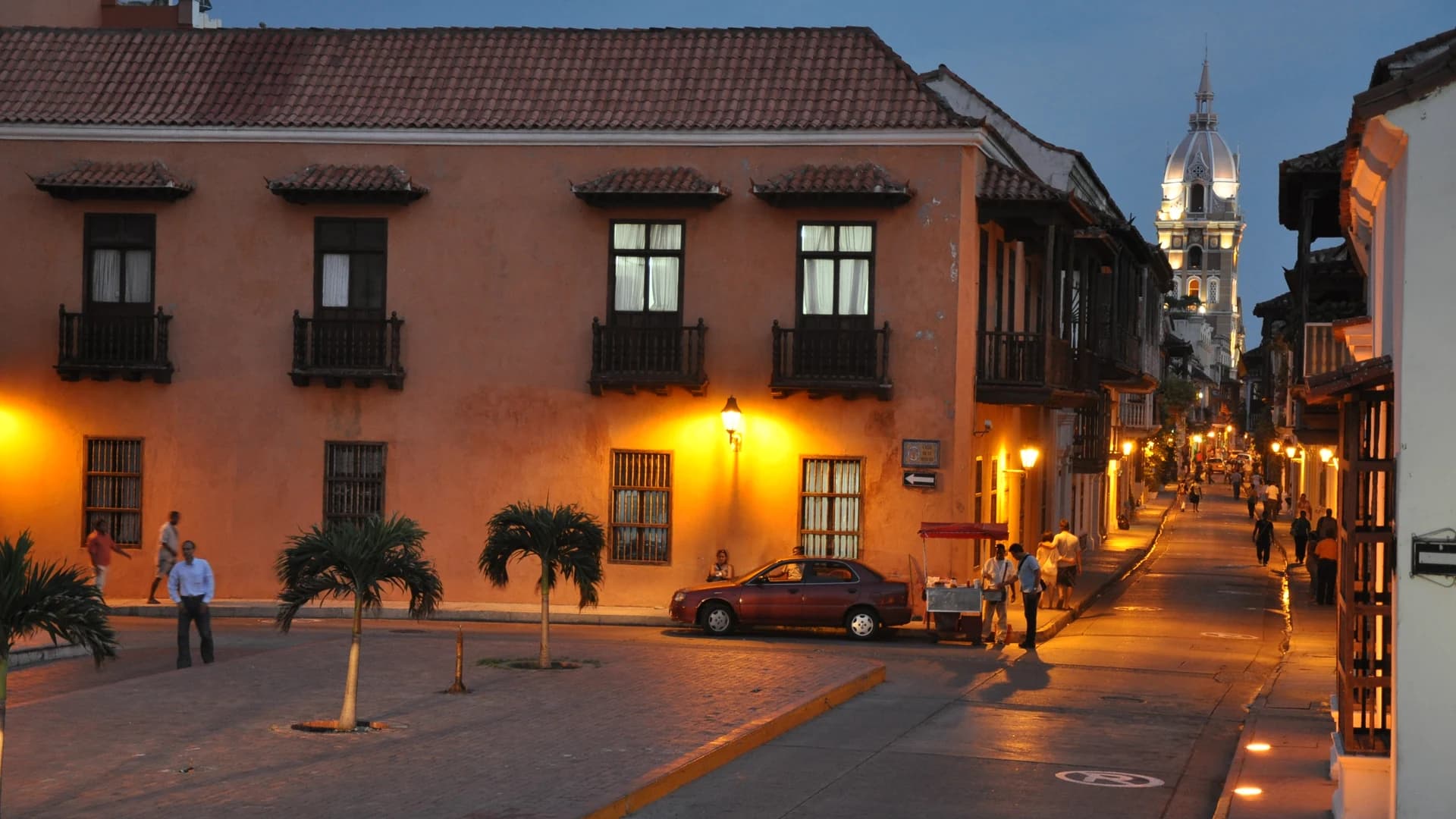 A colonial street at dusk in Cartagena's walled city, with the illuminated cathedral tower in the distance